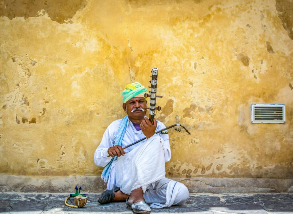 A South Asian musician in traditional attire plays a unique instrument against a rustic wall.