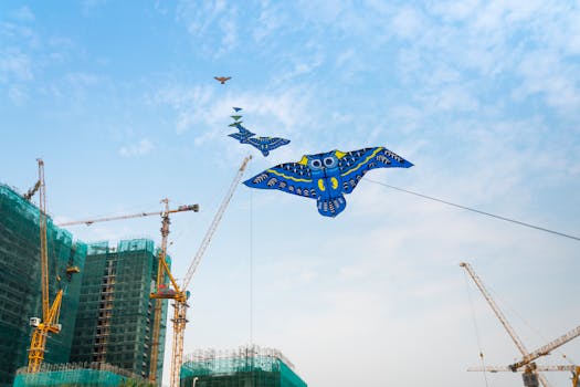 Vibrant owl-shaped kite soaring above cranes at a construction site under blue skies.